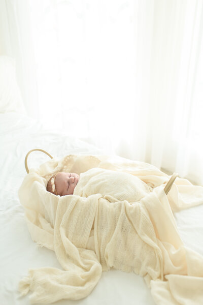 Newborn baby swaddled in cream fabric sleeps peacefully in a basket on a white bed. A dreamy newborn session by Moments in Light Photography.