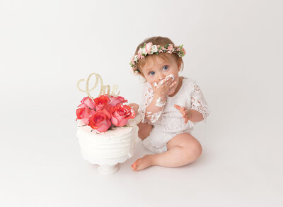Brooklyn, NY Cake Smash Photographer captures baby girl getting messy with her arms covered in cake and eating a handful of icing. Baby is wearing a pink floral crown and fresh flowers top the white cake.