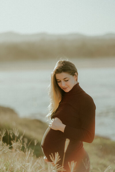 A dreamy maternity photo of a woman holding her pregnancy bump in a field of grass overlooking the beach at Boambee Bay