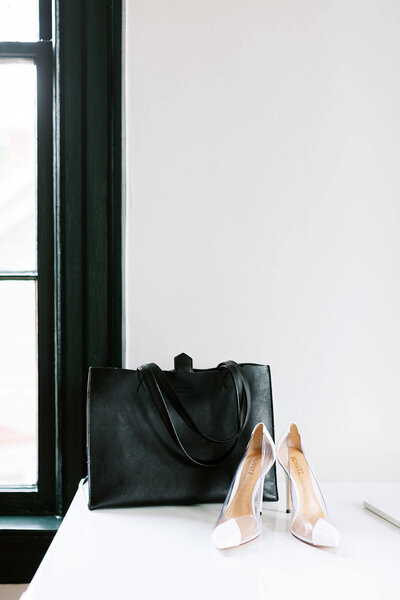 White heels and a black leather bag sitting on a white table