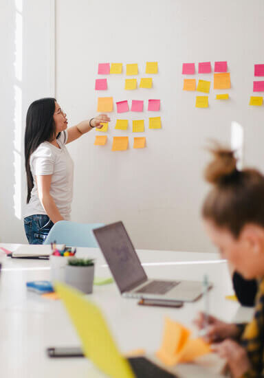 Woman teaching an educational workshop for real estate agents, pointing at a white board with sticky notes in front of a classroom of people