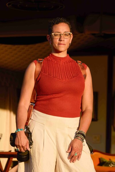 Kate Okenatez-Mahoney, a luxury elopement designer and photographer wears an orange sleeveless top with knit texture and cream colored linen pants, while holding a camera.  She is in front of a luxury safari tent