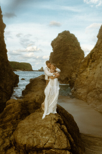 M standing in front of J with their arms wrapped around each other standing on a rock surrounded by the ocean