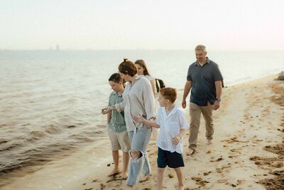 A family of five walks barefoot along a sandy beach near the water on a calm, overcast day.