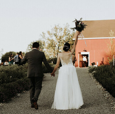 couple walking outside on gravel path lined with greenery and bride raising bouquet overhead