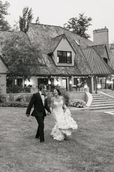 a black and white photo of a bride and groom running down a hill with the venue Basil Place in the background. 