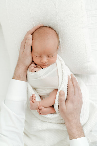 A newborn wrapped in a white blanket while dads hands protects the baby. Captured by Lisa Lefevre Photography