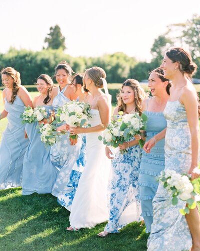 Rhode Island Wedding Photographer | Bridesmaids in blue dresses and a bride in white walk together on a sunny day, holding bouquets, exuding joy and celebration in a lush garden setting.