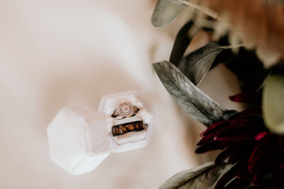 bride and grooms wedding rings in a white box with the brides florals surrounding the m on a white fabric