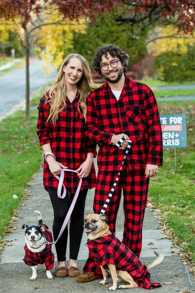 a couple stands in plaid pajamas with their two dogs in matching pajamas