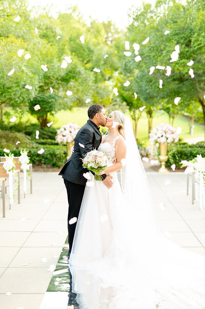Bride and groom kiss as rose petals fall from the sky.