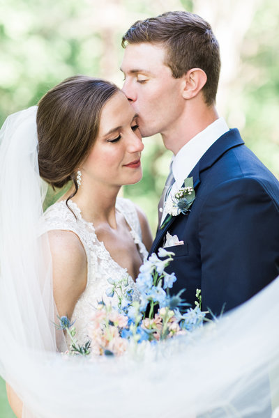 A woman in a white dress with long white veil holding a bouquet being kissed by a handsome man in a navy suit at Silver Hearth Lodge wedding venue.
