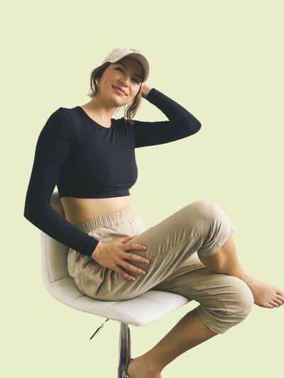 Caitlin sits on a chair with one hand on her cap and a relaxed smile. She wears a black long-sleeve crop top and beige pants against a light green background.