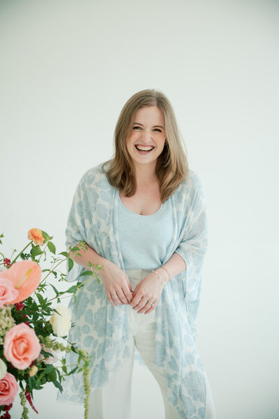Dania, a wedding content creator with long hair sits smiling on a green armchair, wearing a cream top and rust skirt, surrounded by soft pink and white flowers indoors.
