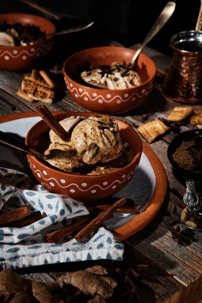 A cozy, rustic image of spiced ice cream served in terracotta bowls, surrounded by cinnamon sticks and other spices. Photographed by Mica McCook, this scene evokes warmth and comfort, perfect for a fall or winter treat.