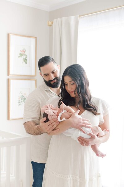 A couple stands in their baby's neutral colored nursery and gazes at their daughter during in-home newborn photos in New Jersey.
