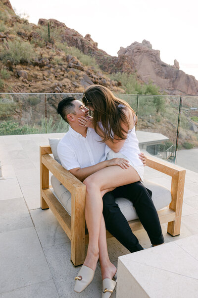 A stylish couple shares a passionate embrace during a golden-hour engagement session in the Scottsdale desert. The bride-to-be, dressed in a chic white outfit, leans into her fiancé with a classic vintage Porsche parked behind them, capturing a timeless and adventurous love story.