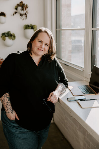 Confident female entrepreneur with floral arm tattoos smiling while standing by a bright office window next to a laptop, phone, and open notebook
