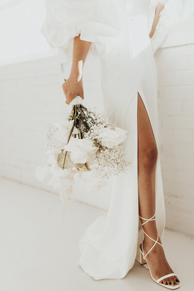 person in white dress and strapy heels standing against a white wall holding a white bouquet