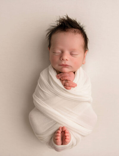 Studio newborn portrait by Stamford CT photographer showcasing peacefully sleeping baby wrapped in cream swaddle against neutral background