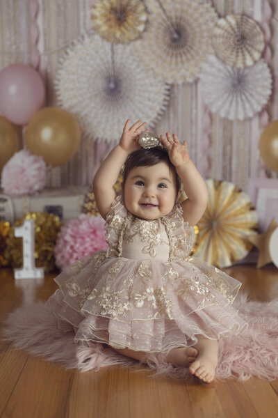 First birthday girl wearing pink & gold dress with matching headband smiling during her studio portrait session.