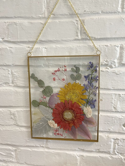 Vivid pressed flower design with bright red and yellow daisies, complemented by eucalyptus and colorful accents in a hanging gold frame against a white brick wall.