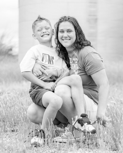 black and white photo of a mom and son in grass.  Mom is knelt down with son on her lap both smiling at the camera.  Son has short spiked hair, wearing shorts, tennis shoes, and a t-shirt.  Mom is wearing shorts, t-shirt, has long hair