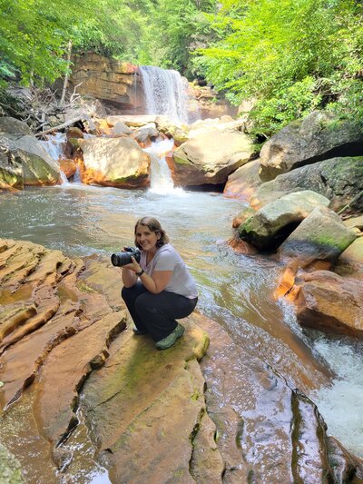 Joy Granados kneeling with her camera in front of a waterfall, behind-the-scenes portrait of Capture Joy Photography, Northern Virginia family photographer.