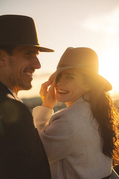 Mountain climb at sunset: Laughing couple wearing Akubra hats sharing a joyful, intimate moment during golden hour, perfect for a relaxed elopement.