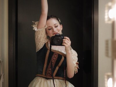 Shelby Shenkman in a period ballet tutu with one arm i nthe air, holding a Nikon camera pointed towards the subject