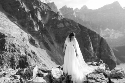 Black and white portrait of a bride playing with her dress and veil on a mountain top at Moraine Lake in Banff National Park during an evening photo session