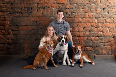 A smiling couple poses in front of a rustic brick wall with their three dogs. The woman sits on the floor with one dog, while the man crouches behind her with the other two dogs sitting in front. The dogs vary in breeds and wear colorful collars, all captured beautifully by a Jacksonville pet photographer.