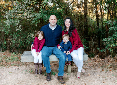 Smiling family of four sitting on a wooden bench in a natural outdoor setting, dressed in coordinating navy, red, and white outfits. Text overlay reads 'Family - View the Details,' highlighting warm and timeless family photography sessions.