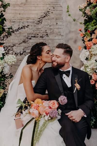 Bride snuggles and kisses groom on cheek while holding bouquet