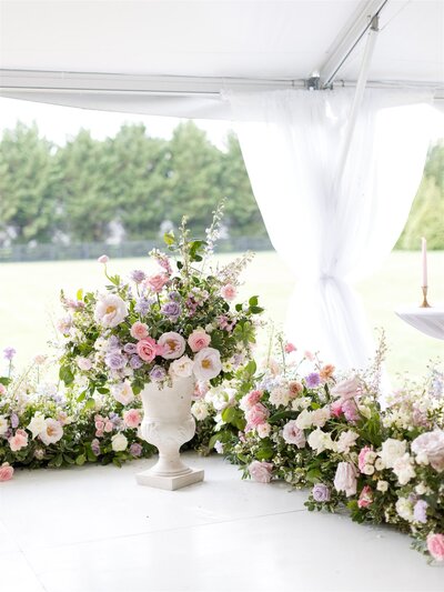 Pastel colored florals line the stage of this summer wedding ceremony with floral urns and meadows. Pink, lavender, cream roses brighten this tented wedding. Design by Rosemary and Finch in Nashville, TN.