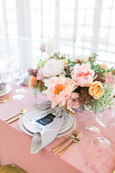 A romantic wedding table set up with a blush pink velvet linen, gold flatware, grey napkin, and white plates. the flowers are large with peach, pink, golden, and burgundy tones. the room has lots of natural light
