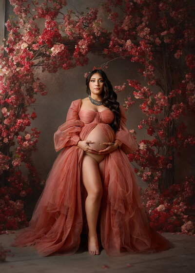 beautiful dark hair woman wearing a soft vintage pink gown and jewelry, showing her belly surrounded by a floral background in pink