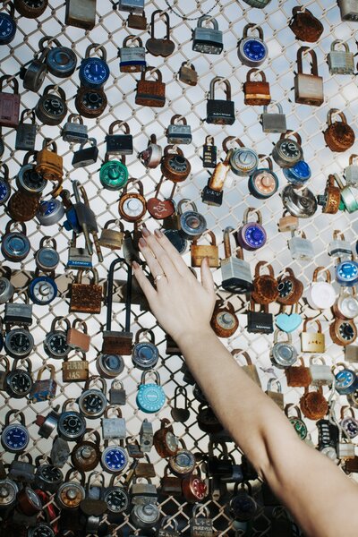 A hand reaches toward a fence covered with colorful locks and combination padlocks, symbolizing love and connection.