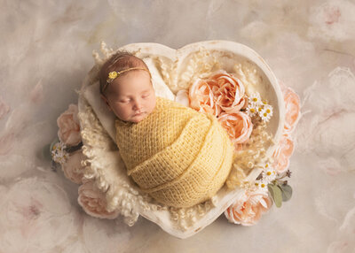 Baby girl at newborn session in heart bowl prop surrounded by pink flowers in a yellow wrap