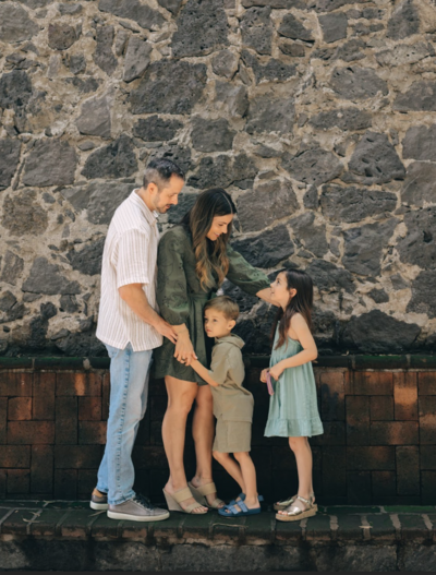 Natalie Bernacchi with her husband and two young children, standing together and smiling in front of a rustic stone wall