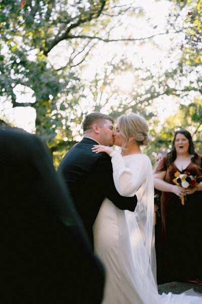 A bride and groom are sharing a kiss and behind them is the maid of honor showing her excitement