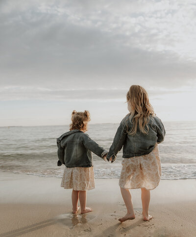Image of two children on a beach, holding hands and facing away from the camera out to the ocean.  