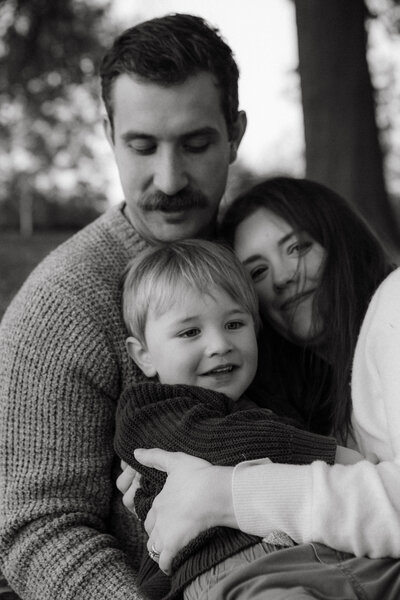 Black and white image of a father holding a mother and their young son, mother smiling at the camera, capturing authentic, emotional family moments that celebrate connection and presence