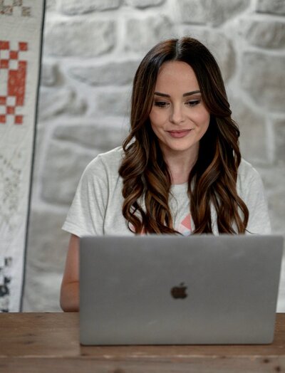 long brown haired girl sitting at a mac laptop with a quilt behind her