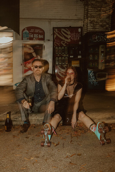 bride and groom sit on sidewalk while sipping champagne and wearing roller skates 