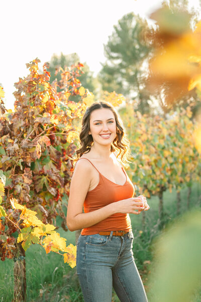 High school senior girl wearing an orange tank top and blue jeans, standing in grape vines in the fall