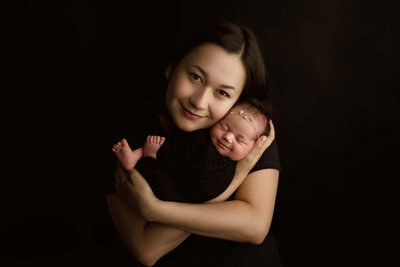 Studio portrait of a mother lovingly holding her newborn baby girl, both dressed in a black against a dark fine art background - a timeless and elegant moment captured at Duehr Photography in Maple Grove,Minnesota