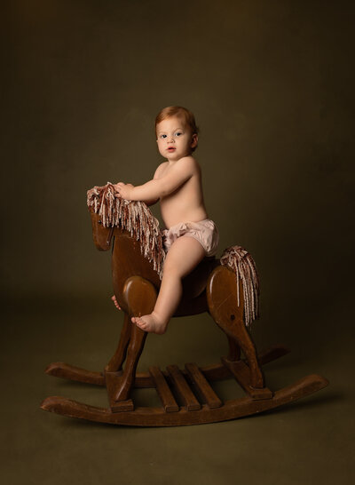First birthday photoshoot. Baby boy sitting on a wood rocking horse wearing just a diaper cover. He is looking over his shoulder to the camera. The rocking horse is on a olive green backdrop. Captured by best Brooklyn, NY milestone photographer Chaya Bornstein.