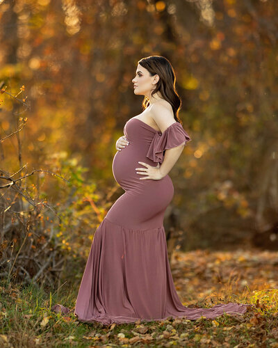 A beautiful pregnant woman in a mauve dress is standing in a grassy area with the sun creating a halo around her