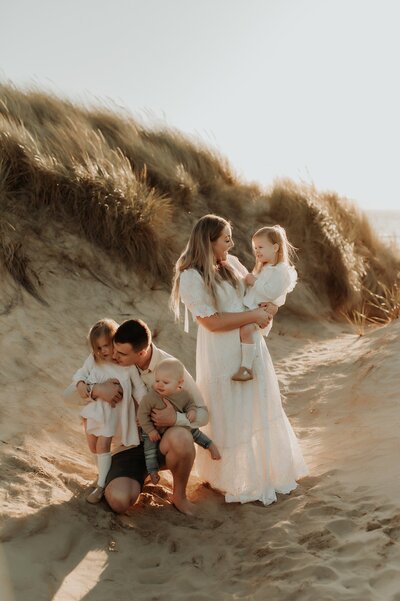 Young family with 3 little ones plays on a beach for candid family photos. Mom is wearing a long dress and everyone is in neutral colors